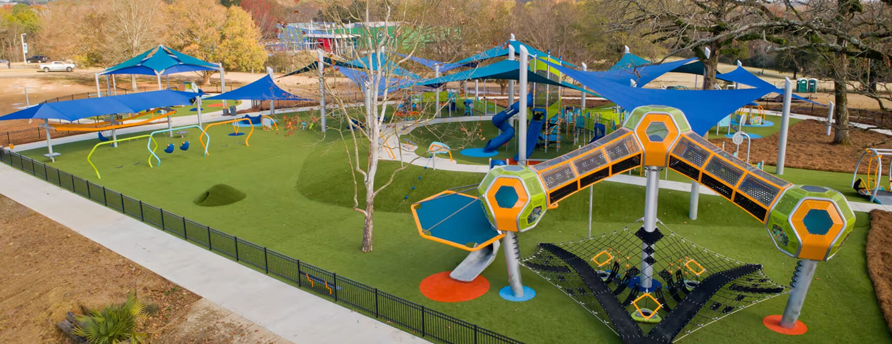 Modern playground featuring climbing structures, slides, swings, and blue shade canopies on green turf.