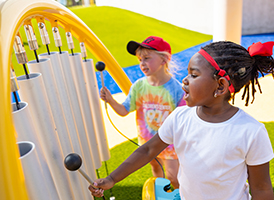 Two young kids play outdoor chimes side by side on a sunny day, smiling as they make music together.