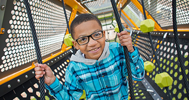 Boy with glasses smiles as he climbs a playground structure made of ropes and green geometric shapes.