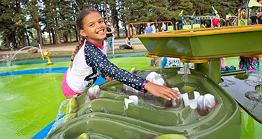 A smiling girl plays in a splash park, splashing water with trees and other children in the background.