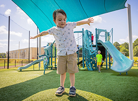 Boy balances with arms outstretched on playground turf; slides and a shade canopy are visible behind him.